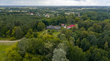 Polonya 'nın Nowy Lubiel kentindeki ormanların, tarlaların ve dağınık binaların karışımı. Parçalı bulutlu bir gökyüzünün altında kırmızı çatılı bir ev, toprak yol ve tarım arazisi bulunuyor..