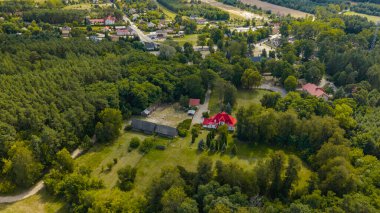 Polonya 'nın Nowy Lubiel kentindeki kırmızı çatılı evin hava görüntüsü, yeşillikler, ağaçlar, çiftlik yapıları, sık ormanlar ve uzak bir kırsal kasaba ile çevrili..