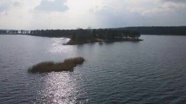 Sakin bir gölde yapraksız ve yemyeşil ağaçlarla dolu küçük bir adanın havadan görünüşü. İHA sürekli hareket ederken güneş ışığı suya yansıyor..