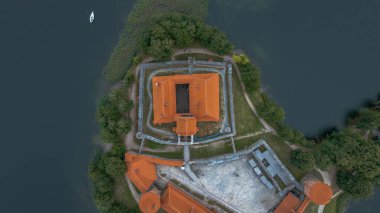 Kırmızı turuncu çatıları olan Trakai Adası Kalesi, yeşil ve suyla çevrili Galve Gölü 'nde bulunur. Durgun gölde beyaz bir tekne görünüyor..