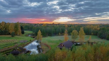 Gün batımında sakin Litvanya manzarası, bir gölet, ahşap evler, sonbahar ağaçları, sık orman ve turuncu ve pembe renkli canlı bir gökyüzü.