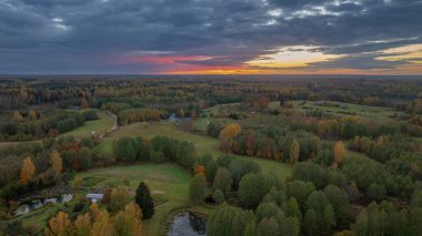 Günbatımında Litvanya 'nın sakin kırsal manzarası, sonbahar yeşillikleri, tarlalar, ormanlar, kırsal evler, göletler ve dramatik aydınlatılmış bulutlar.