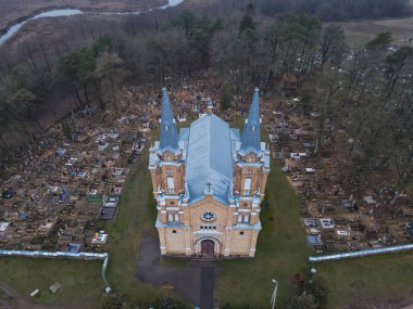 Açık renkli tuğlaları, ikiz kuleleri ve mavi çatısı olan bir Katolik kilisesinin hava manzarası. Etrafı mezarlık, ağaçlar ve arka planda bir nehir ile çevrili..
