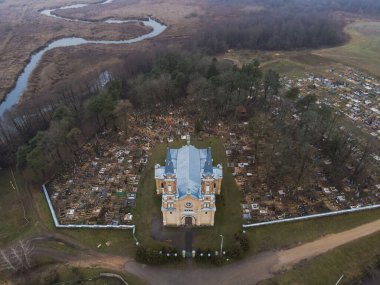Açık renkli, mavi çatılı bir Katolik kilisesi ağaçlarla, açık arazilerle ve arka planda dolambaçlı bir nehirle çevrili bir mezarlığın yanında duruyor..