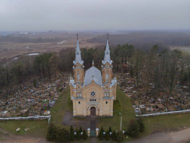 İkiz kuleleri ve ön cephesi olan bir Katolik kilisesi bir mezarlığın ortasında duruyor, ağaçlarla çevrili ve bulutlu bir gökyüzünün altında açık araziler..
