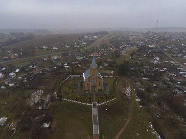 Yüksek çatısı ve yüksek kulesi olan bir Katolik kilisesi, bulutlu bir gökyüzünün altında beyaz çitlerle çevrili dağınık evler, tarlalar ve kar parçalarıyla çevrili bir tepenin üzerinde duruyor..