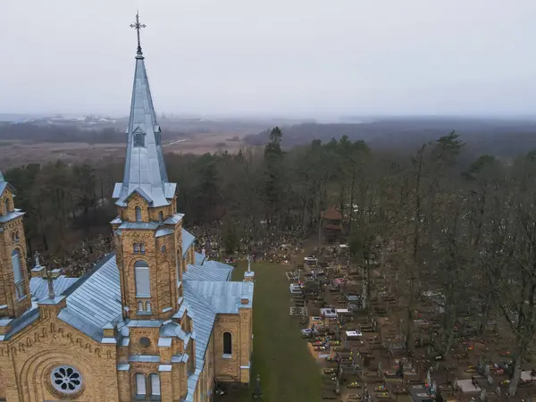Mavi metal çatılı ve yüksek kuleli tuğla bir Katolik kilisesi, Belarus 'ta bulutlu bir gökyüzünün altında orman ve tarlalarla çevrili bir mezarlığın yanında duruyor..