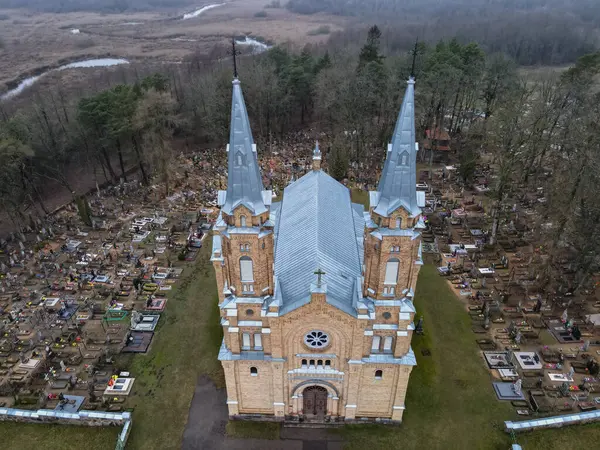 Sivri kuleleri olan tuğladan bir Katolik kilisesi mavi metal bir çatı ve etrafı mezarlık, ağaçlar ve su yolu olan bataklık manzarasıyla çevrili bir gül penceresi..