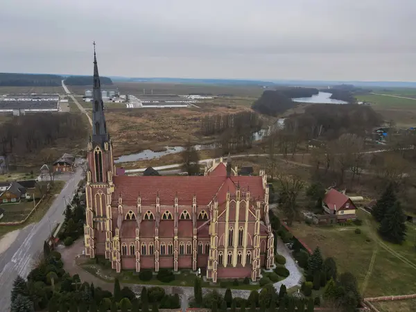 Beyaz Rusya kırsalında, bahçeler, dağınık evler, tarlalar, bir nehir ve bulutlu gökyüzü ile çevrili yüksek kuleli neo Gotik bir Katolik kilisesi bulunuyor..