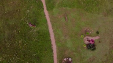 Toprak yolu olan yeşil bir tarlanın havadan görünüşü, ortaçağ giysileri içindeki iki birey ve küçük beyaz çiçekler. Kamera yavaş yavaş döner ya da döner..