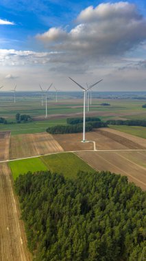 Tarımsal alanlarda rüzgar türbinlerinin manzaralı görüntüsü, önplanda yoğun bir orman ve kırsal sürdürülebilirliği vurgulayan parçalı bulutlu bir gökyüzü..