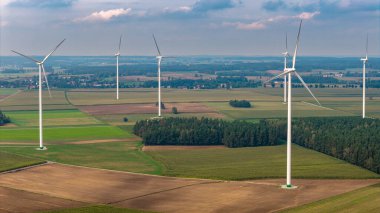 Rüzgâr türbinleri, ağaç kümeleri ve parçalı bulutlu gökyüzünün altındaki küçük yerleşim alanları kırsal bölgelerde yenilenebilir enerji sergiliyor..