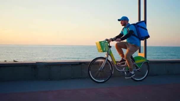 青い制服を着たポジティブな配達人が自転車や日没や日の出に乗って — ストック動画