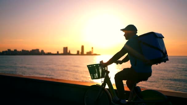 バックパックを背負った配達員が自転車に乗って街の背景を背景に日没時に — ストック動画