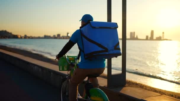 身着蓝色制服 背着食物背包的送货员日落时在一座现代化城市的背景下散步 从后面看风景 — 图库视频影像