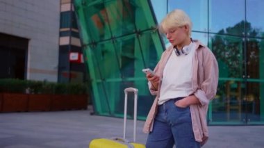 Genç ve çekici bir bayan gezgin, kulaklıkları ve elinde akıllı bir telefonla modern bir binanın arka planına bakarak.