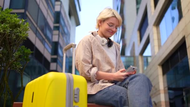 スーツケースを持った若い女性旅行者が座ってスマホを使って笑っています — ストック動画