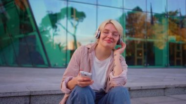 Basamaklarda oturan, kulaklıklı ve elinde telefon olan genç ve pozitif bir kadın.