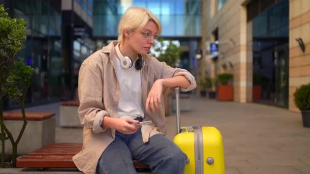 スーツケースを突き刺すような非公式な外観の若い女性旅行者が座っている 混乱しているか悲しい — ストック動画