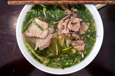 Vietnamca pho bo baharatlı çorba ya da beyaz seramik kasede pirinç eriştesi, sığır eti dilimleri ve ahşap arka planda bitkiler. Kuzey Vietnam halkının zevkine ve tarzına göre pişirildi.