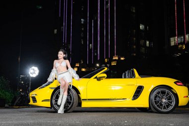 Hanoi, Vietnam - 26 Haziran 2024: Porsche 718 Boxster ile bikini giyen güzel genç bir kadın moda fotoshoot sırasında sokağa park etmiş. Porsche, 1931 'de kurulan yüksek performanslı spor arabalar konusunda uzman bir Alman otomobil üreticisi..