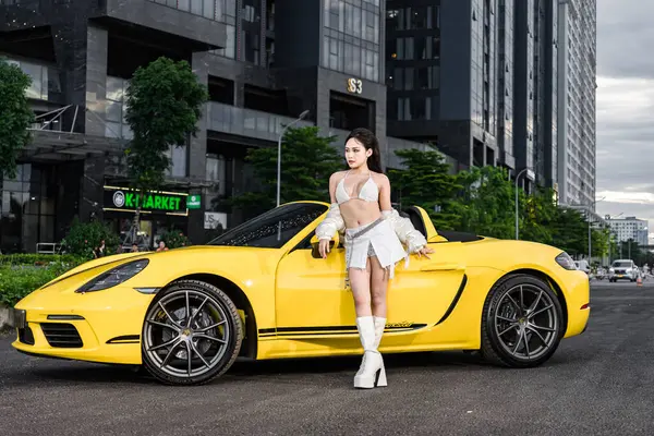 Hanoi, Vietnam - 26 Haziran 2024: Porsche 718 Boxster ile bikini giyen güzel genç bir kadın moda fotoshoot sırasında sokağa park etmiş. Porsche, 1931 'de kurulan yüksek performanslı spor arabalar konusunda uzman bir Alman otomobil üreticisi..