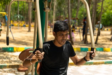 Genç bir adam hareketli bir çocuk parkında egzersiz yaparken terler. Yeşillik ve oyun alanı ekipmanlarıyla güç, dayanıklılık ve esneklik kazanmak için çeşitli egzersizler yapıyor. Faydalarını ilk elden gör.