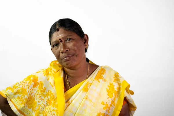 A South Indian village woman with a sad face looks directly at the camera. She is wearing a traditional yellow saree and has her hair tied back