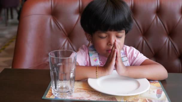 彼女の前でピンクのドレスとプレートとガラスを待っているレストランで食べ物の前に祈る子供のビデオです — ストック動画