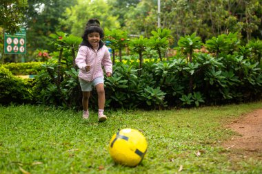 Futbol formalı genç bir kız parkta koşup futbol topunu tekmeliyor. Gülümsüyor ve eğleniyor. Güneş parlıyor..