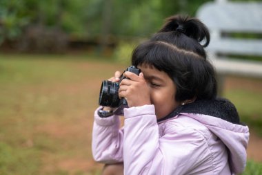 Genç bir kız, yaklaşık 6 yaşında, elinde bir DSLR kamerasıyla fotoğraf çekiyor. Pembe bir elbise giyiyor..