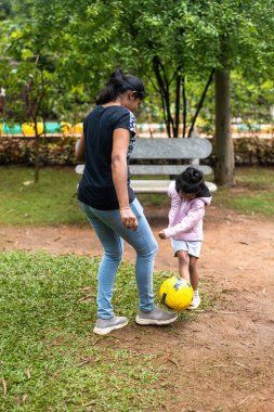 5 yaşındaki mutlu bir kız ve annesi parkta futbol oynuyorlar. Kız gülüyor ve topun peşinden koşuyor.