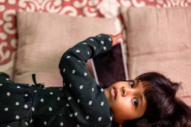 Tablet 'in mobil bağımlılık konsepti ile koltukta uzanan sevimli küçük bir kız.