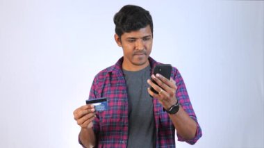20 'li yaşlarda genç bir adam bir elinde cep telefonu, diğer elinde kredi kartı tutuyor. Gülümsüyor ve telefon ekranına bakıyor.,