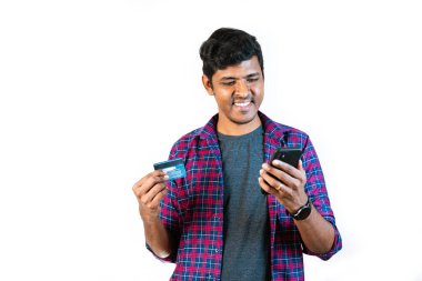 20 'li yaşlarda genç bir adam sağ elinde bir cep telefonu ve sol elinde bir kredi kartı tutuyor. Bir ödeme uygulaması gösteren telefon ekranına bakıyor.