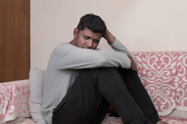 Genç bir Hintli adam oturma odasındaki bir kanepede tek başına oturuyor, derin düşünceler içinde. Onun ifadesi tefekkür ve üzüntünün bir karışımıdır.