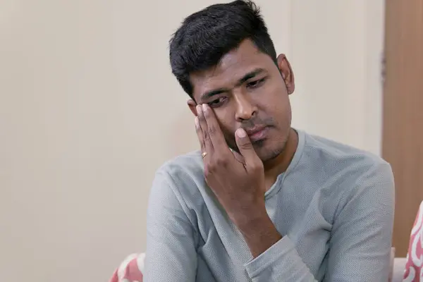 stock image A young South Asian man, seated on a sofa at home, wipes away tears while gazing directly at the camera