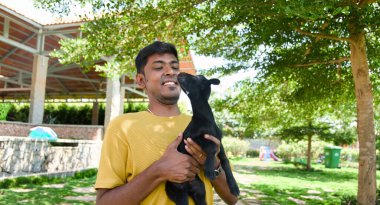 Sıcak bir gülümsemesi olan kibar bir Hintli güzel bir parkta güneşli bir günde kollarında minik siyah bir keçi beşiği taşıyor..