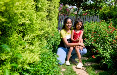 Canlı bir bahçede birlikte oturan, kameraya doğru gülümseyen bir anne ve kızının yürek ısıtan görüntüsü.