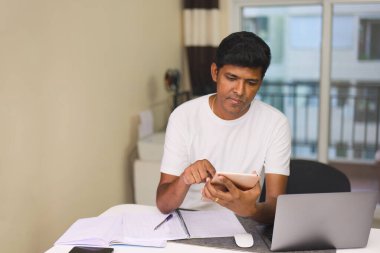 Genç Hintli bir adam, oturma odasında hem laptop hem de dijital tablet kullanarak verimli bir şekilde çalışır.