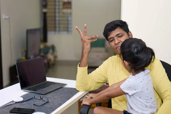 Stock image A warm and inviting scene of a caring Indian father guiding his young daughter as she explores the world of computers on a laptop at their home desk.