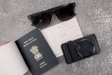 Bu resim seyahat gereksinimlerini gösteriyor: düz gri bir taş yüzeyde düzenlenmiş bir Hindistan pasaportu, kamera, güneş gözlüğü ve bir çek..