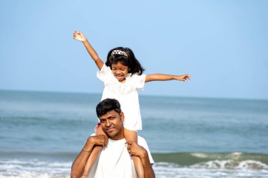 Bir baba kızını omuzlarında taşır, her ikisi de okyanus zeminine gülümserler. Sevgi ve oyun dolu bir an..