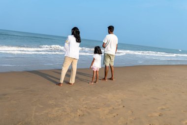 Beyaz giyinmiş üç kişilik bir aile, okyanus dalgalarının kıyısında huzurlu bir anın tadını çıkarıyor.