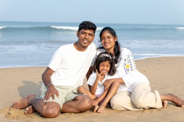 Beyaz giyinmiş üç kişilik bir aile, okyanus dalgalarının kıyısında huzurlu bir anın tadını çıkarıyor.