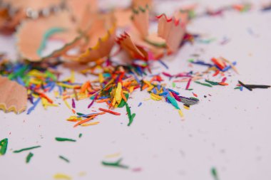 Beyaz bir yüzeye dağılmış renkli kalem talaşlarının yakın plan görüntüsü. Talaşlar çeşitli renkli kalemlerden ve rastgele dizilmiş desenler. Bazı talaşlar hala kaleme bağlıyken, diğerleri gevşek ve dağınık.