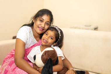 Bir anne ve bir çocuk, aralarında doldurulmuş bir köpekle koltukta oturuyorlar. Kadın pembe bir sari giyiyor ve çocuk beyaz bir gömlek giyiyor. Sahne sıcak ve sevgi dolu, tıpkı bir kadın gibi.