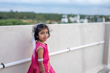 Renkli geleneksel Hint kıyafetleri içinde genç bir kız çatıda duruyor. Parlak bir gülümsemeyle manzaranın huzurlu manzarasının tadını çıkarıyor..