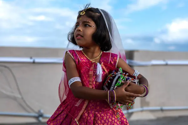 stock image A young girl dressed in vibrant traditional clothing stands confidently, holding colorful musical instruments while participating in a joyous cultural celebration under a clear blue sky.