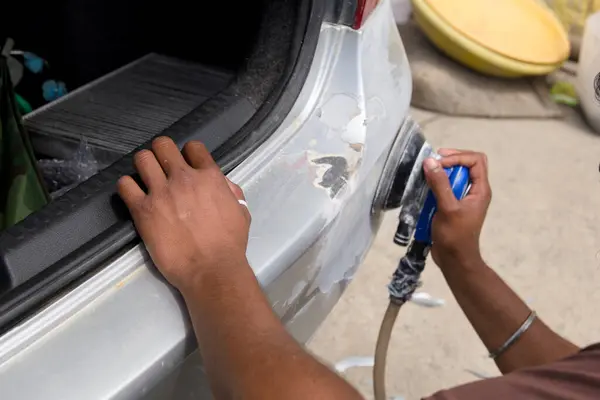 stock image A technician works on a vehicle's rear, applying body filler to smooth out imperfections before paintwork. The setting is a busy auto shop filled with tools and materials.